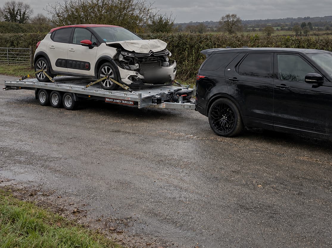 A white hatchback with front-end crash damage strapped onto a car trailer